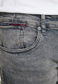 Harmaat farkut klassisella designilla, jossa on kolikoita varten tasku punaisella ja sinisellä ompeleella sekä brändillä varustettu metallipainike vyötäröllä.