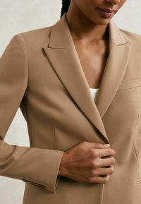 Smėlio spalvos švarkas su pritaikyta siuvimo konstrukcija, struktūruotais atvartais ir viena priekine kišene. Lygi medžiaga su subtiliu blizgesiu ir rankogaliais su sagomis.