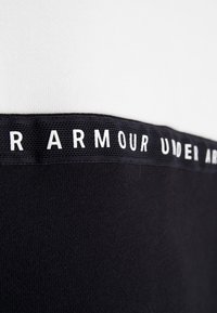 Camisa de manga corta con parte superior blanca y parte inferior negra, que presenta una banda texturizada negra con "UNDER ARMOUR" en letras blancas.