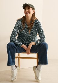 Groene geblokte blouse met lange mouwen en kraag, gecombineerd met blauwe wijde jeans en witte sneakers. Zittend op een houten stoel.