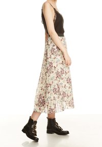 Donna che indossa un top nero con pizzo, una gonna midi plissettata crema a fiori e stivaletti neri alla caviglia con fibbie dorate, che cammina verso destra.