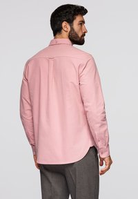 Homme aux cheveux foncés et barbe portant une chemise rose à manches longues et un pantalon gris, de profil avec principalement le dos visible.