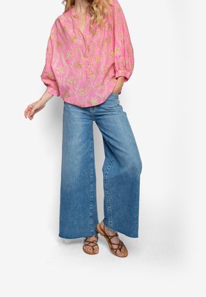 Kvinde iført løs lyserød bluse med guld palmeprint, blå jeans med vide ben og brune sandaler med stropper, stående mod hvid baggrund.