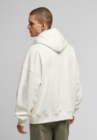 Hvid oversized hættetrøje med en stor hætte, ribbede manchetter og kant. Blød tekstur, sømløse skuldre og en afslappet pasform, vist fra bagsiden.