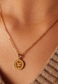 Gouden hangerketting met een ronde charme in klaverontwerp; schakelkettinkje met een gladde afwerking, rustend op de huid.