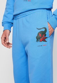 Blue sweatpants with elastic waist, featuring a green lizard with red flower graphic and Lacoste and Netflix logos near the right pocket.