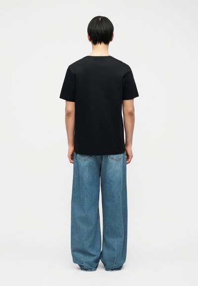 Άτομο με την πλάτη προς τα έξω φοράει μαύρο κοντομάνικο πουκάμισο και φαρδιά μπλε τζιν, σε απλό φόντο.