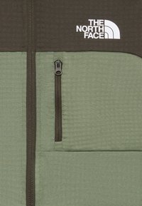 Veste verte et marron avec des zips verticaux et le logo blanc "The North Face" sur la partie supérieure droite de la poitrine.