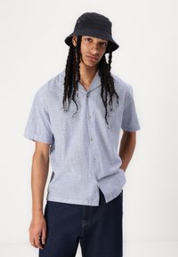 Cămașă cu mânecă scurtă, cu dungi verticale albastre și albe, guler cu nasturi și croială lejeră. Asortată cu blugi negri din denim și o pălărie tip bucket neagră.