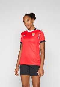 Rød idretts skjorte med svarte detaljer og hvit kant, med Benfica-logoen. Kombinert med svarte shorts, laget av glatt, lett stoff.