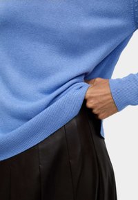 Ljusblå stickad tröja med ribbade muddar och avslut, lagd över en mörk plisserad läderskjorta. Texturen verkar mjuk och slät.