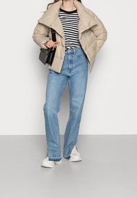 Beżowa kurtka puchowa z wysokim kołnierzem, czarno-biała koszula z długim rękawem w paski, jasnoniebieskie jeansy o prostym kroju oraz beżowe trampki z teksturowaną podeszwą.