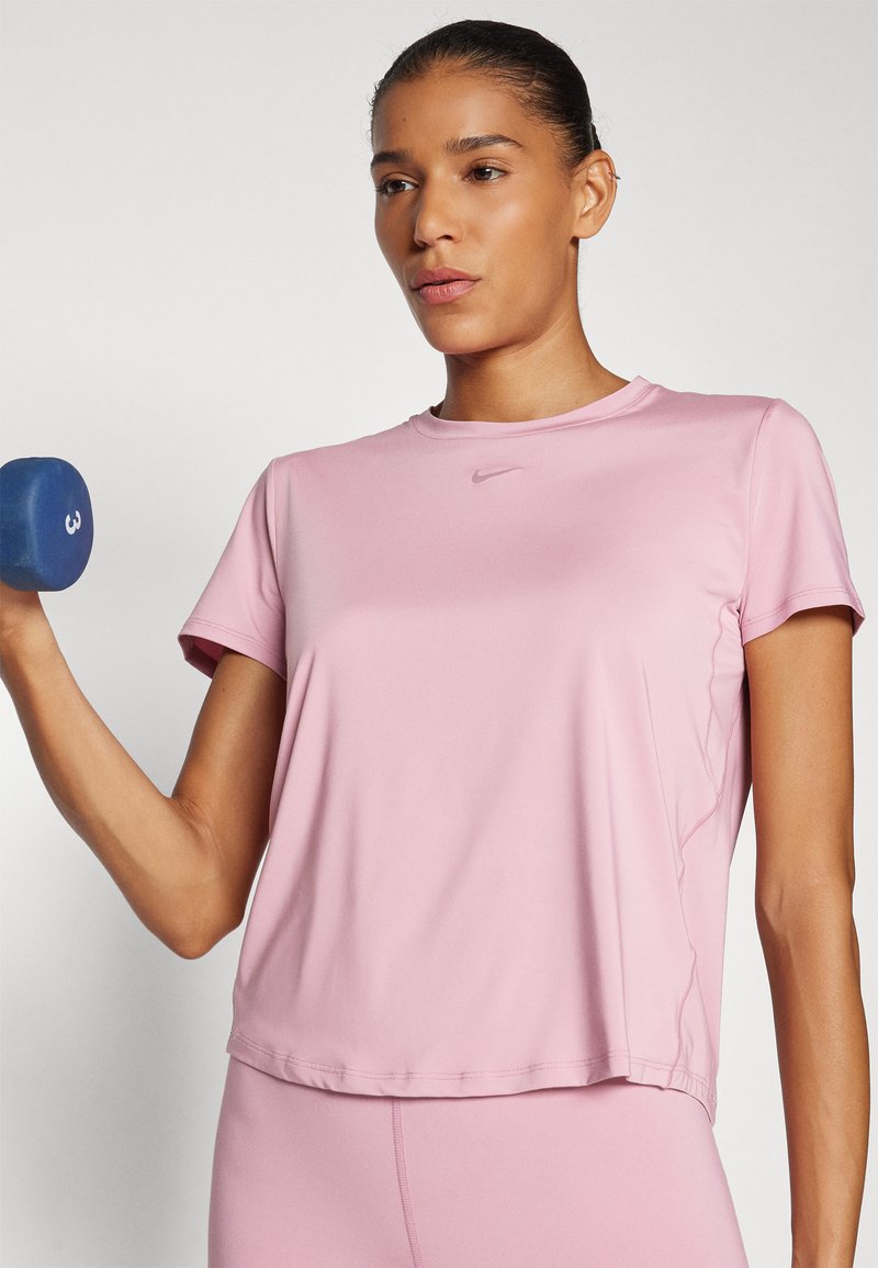 Rosa idrottströja med korta ärmar. Slät tyg, rund halsringning och Nike-logotyp på bröstet. Modellen håller en blå hantel.