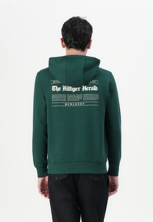 Sudadera verde oscura con un bolsillo canguro, que presenta un gráfico impreso en la parte posterior. El diseño incluye texto y bordes decorativos.