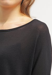 Czarny, lekki sweter z dzianiny o szerokim dekolcie i teksturowanej tkaninie, spoczywający tuż nad ramionami. Gładki i półprzezroczysty wygląd.