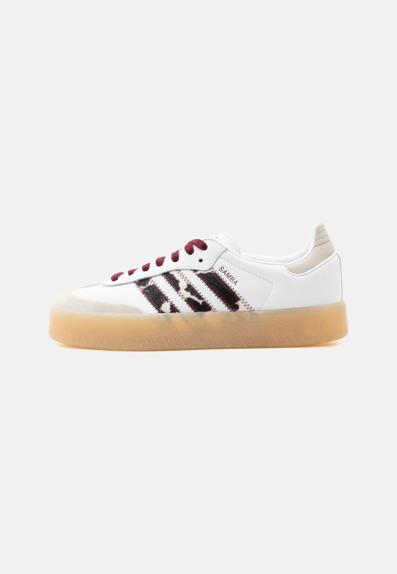 Λευκό αθλητικό παπούτσι Adidas Samba με σκούρες κόκκινες κορδόνια, μαύρες χνουδωτές ρίγες, μπεζ ταμπελάκι στη φτέρνα και διαφανές καουτσούκ σε σόλα, εμφανιζόμενο από αριστερή πλευρά.