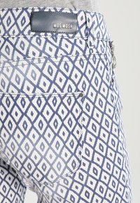 Pantalones de tela blanca con un patrón de diamantes azules, con una etiqueta de logo en la cintura y dos bolsillos traseros. Apariencia texturizada.