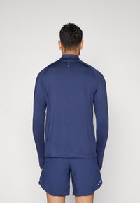 Μπλε ναυτικού χρώματος αθλητικό μπλουζάκι με μακριά μανίκια, ψηλό γιακά και αντανακλαστική λεπτομέρεια στην πλάτη, σε συνδυασμό με ταιριαστά σορτς μπλε ναυτικού χρώματος. Ομαλή υφή υφάσματος.