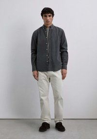 Camisa gris de botones con botones blancos, combinada con pantalones color crema. La camisa tiene cuello y un ajuste relajado. Zapatos marrones completan el look.