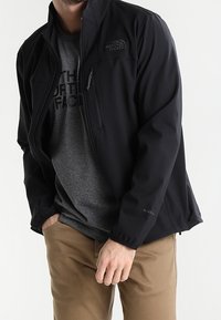 Schwarze Softshelljacke mit hohem Kragen, Reißverschluss-Brusttasche und elastischen Bündchen. Kombiniert mit einem grauen T-Shirt und khakifarbenen Hosen.