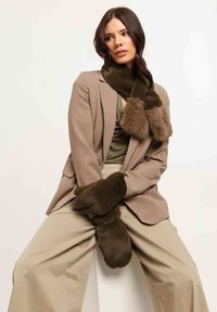 Sciarpa in pelo sintetico verde oliva con due estremità morbide, abbinata a guanti coordinati. Il modello indossa un blazer beige e pantaloni larghi beige.