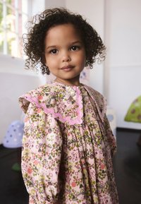 Šaty s kvetinovým vzorom v ružovej farbe s nafúknutými dlhými rukávmi, volánovým golierom a zapínaním na gombíky. Obsahujú zelené, červené a modré akcenty.