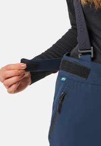 Eine Hand passt eine marineblaue Hose mit einem Klettverschlussbund und einer Reißverschlusstasche an, die ein kleines schwedisches Flaggenetikett und schwarze Hosenträger hat.