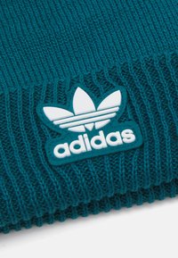 Tirkīza adīta cepure ar ribotu tekstūru un baltu gumijas Adidas zīmi ar trim svītrām un lapu dizainu.