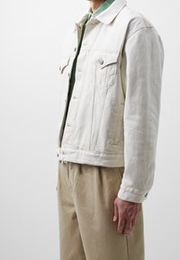 Mand iført en off-white denimjakke over en grøn skjorte og beige bukser, står mod en ensfarvet lys baggrund.
