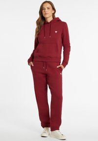 Vrouw staat met handen in de zakken, gekleed in een bijpassende bordeauxrode hoodie en joggingbroek, met witte high-top sneakers, op een witte achtergrond.