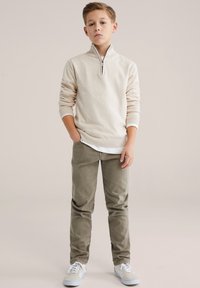 Lysebeige sweater med lynlås-hals over en hvid skjorte, kombineret med olivengrønne bukser og lysegrå sneakers. Enkel strikket tekstur.