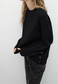 Zwarte gebreide trui met lange mouwen, voorzien van geribde manchetten en knoopaccenten aan de zijkant. Gecombineerd met zwarte leren broek.