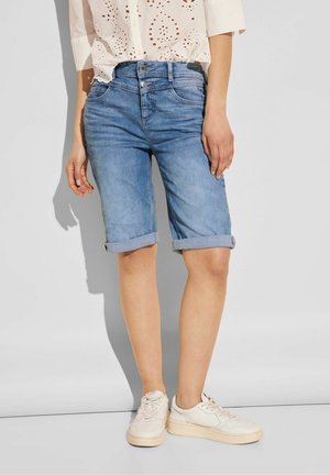 Helle blaue Jeansshorts mit umgeschlagenen Säumen, verziert mit Stickdetails und einem Standardbund. Kombiniert mit weißen Schnürschuhen.
