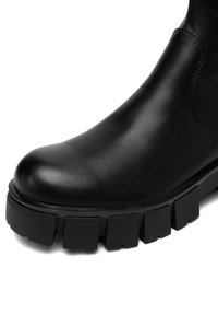 Czarny skórzany botek za kostkę z zaokrąglonym noskiem, masywną teksturowaną podeszwą i elastycznymi panelami po bokach, ułatwiającymi zakładanie.