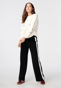 Crème trui met zwarte hartpatronen, gecombineerd met zwarte broek met witte zijstrepen en bordeauxrode sneakers. Soepele textuur, relaxte pasvorm.