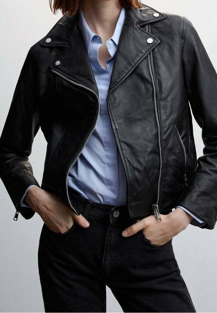 Personne portant une veste de motard en cuir noir sur une chemise bleue claire boutonnée, les mains glissées dans les poches d'un jean noir.