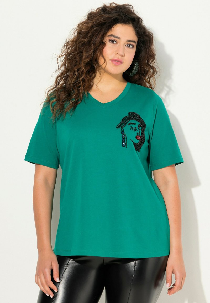 T-shirt verde a V realizzato in cotone, con un design di un volto realizzato con perline nere e colorate sul davanti. Maniche corte, vestibilità rilassata.