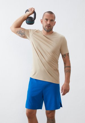 Bežové sportovní tričko s krátkým rukávem, v kombinaci s modrými šortkami. Osoba drží černou kettlebell a na ruce jsou viditelné tetování.
