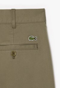 Pantalon khaki beige avec une texture lisse, présentant une poche arrière, un logo crocodile vert et une fermeture à bouton.