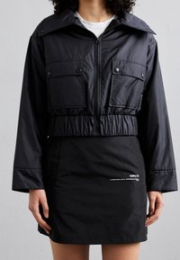 Černá zkrácená bunda se širokým límcem a dvěma předními kapsami; doplněná černou sukní s bílým nápisem na lemu. Hladká, voděodolná látka.