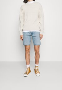 Persona que lleva un suéter crema, una camiseta blanca debajo, pantalones cortos de mezclilla azul claro, calcetines blancos y zapatillas de lona altas mostaza.