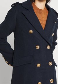 Donna che indossa un cappotto doppiopetto blu navy con bottoni dorati ornate sopra un maglione marrone testurizzato, posata con le mani dietro la schiena.
