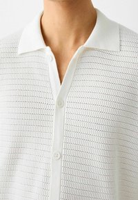 Polo blanco de manga corta con un patrón de tejido texturizado, que presenta un cuello estándar y tres botones blancos en la parte delantera.