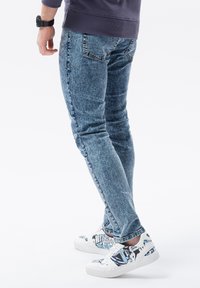 Bleka blå slim-fit jeans med en strukturerad yta, kombinerat med vita sneakers med en färgglad grafisk design och svarta detaljer.