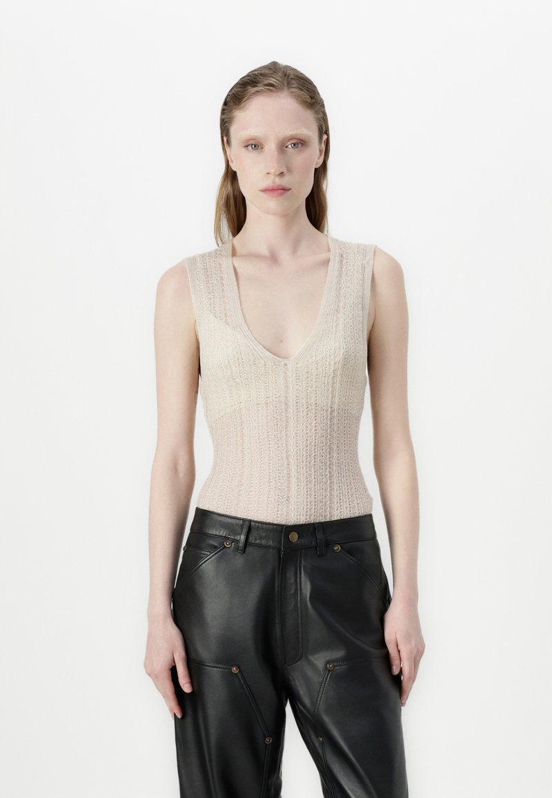 Bež pletena brezrokavnica z globokim V-izrezom, v kombinaciji z črnimi usnjenimi hlačami z okrasnim šivom in gumbi.