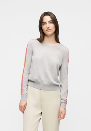 Femme portant un pull léger gris avec des rayures roses et jaunes sur les manches et un pantalon de couleur crème, debout devant un fond uni.