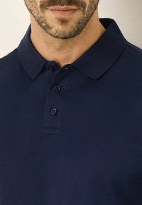 Mörkblå polo med klassisk krage, treknappsslutning och slät textur. Har ribbad krage och fin sömnad.