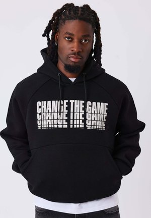 Mann mit geflochtenem Haar, der einen schwarzen Hoodie mit weißem Text "CHANGE THE GAME" trägt, steht vor einem schlichten hellen Hintergrund.