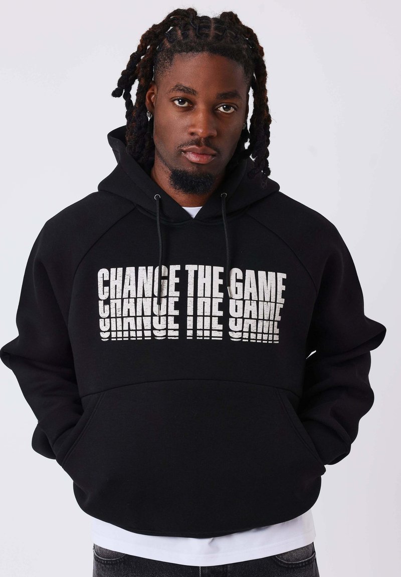 Homme aux cheveux tressés portant un sweat à capuche noir avec un texte blanc "CHANGE THE GAME", debout devant un fond clair uni.
