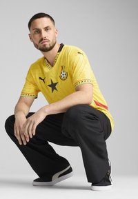 Žltý futbalový dres so starým a futbalovou grafikou, krátke rukávy, čierne akcenty na golieri a rukávoch, spárovaný s čiernymi nohavicami a teniskami.